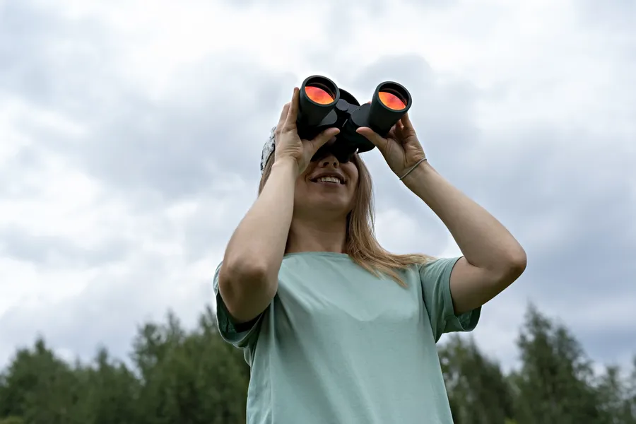 Nuori nainen hymyilee käyttäessään kiikareita, katsoen taivasta kohti pilvisten sääolojen aikana. Hänellä on päällään vaaleansininen t-paita ja pitkä vaaleahiuksinen kampaus.