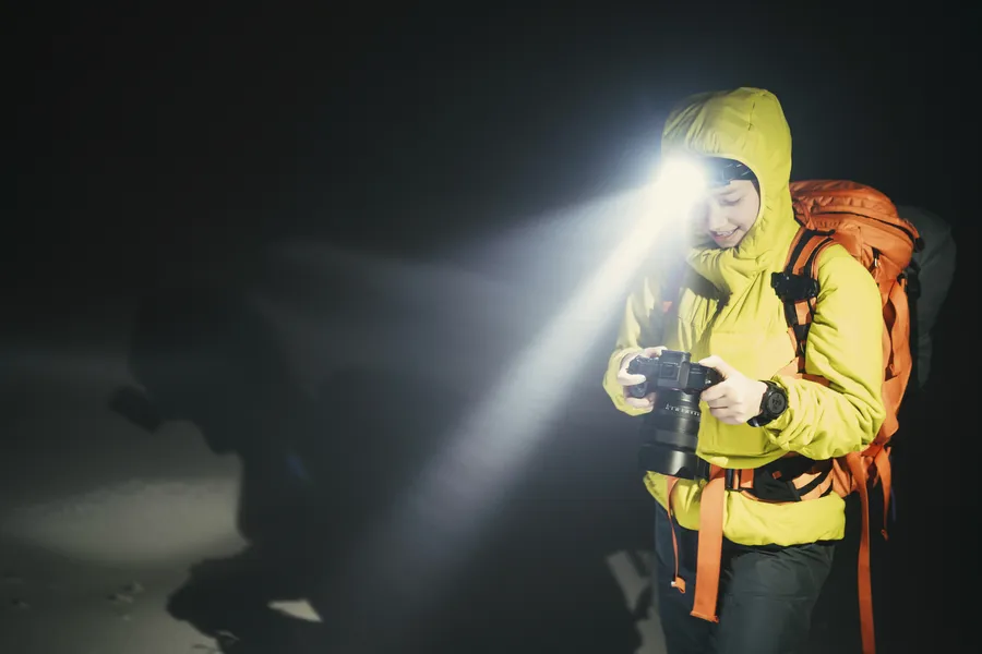 Nuori vaeltaja keltaisessa takissa käyttää otsalamppua ja kuvaa pimeässä metsässä. Taustalla näkyy varjoja ja valoa.