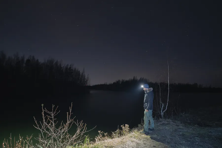 Mies seisoo pimeässä metsässä otsalamppu päässään, tutkien ympäristöä veden äärellä.
