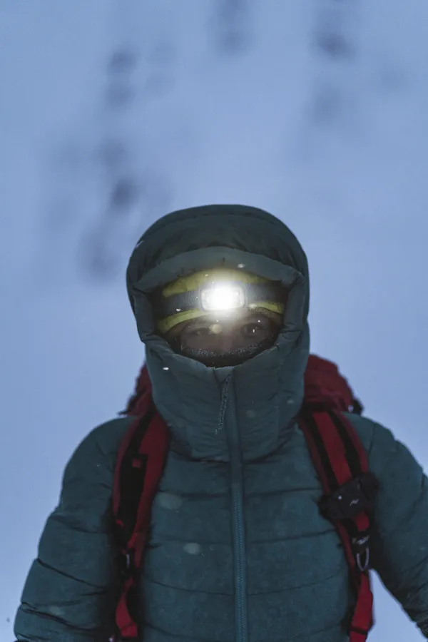Metsävaeltaja pukeutunut haalarinomaiseen takkiin, käyttää kirkasta otsalamppua lumisella polulla.