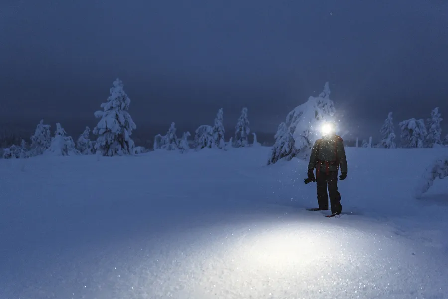 Mies lumisessa maisemassa käyttää otsalamppua valaisten tietään pimeässä talvi-illassa.