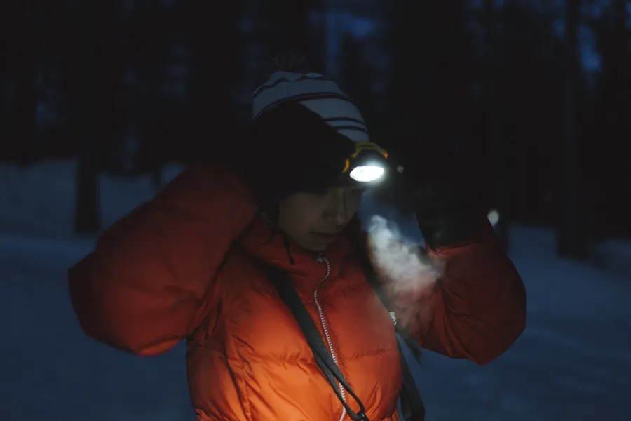 Lumisen metsän äärellä oleva nainen säätää otsalamppua tummassa talvivaatteessa.