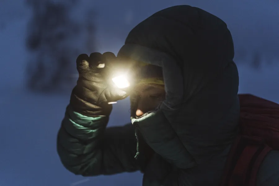 Mies talvivaatteissa käyttää otsalamppua pimeässä metsässä, etsien suuntaa lumisissa olosuhteissa.