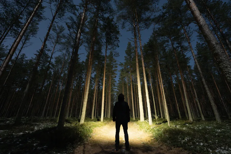 Mies seisoo pimeässä metsässä, käyttää otsalamppua valaisten polkua ja ympäröivää luontoa.