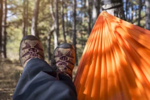 Miehen jalat riippumatossa orange värisessä riippumatossa metsän keskellä, taustalla puita ja luonnon rauhallisuus.