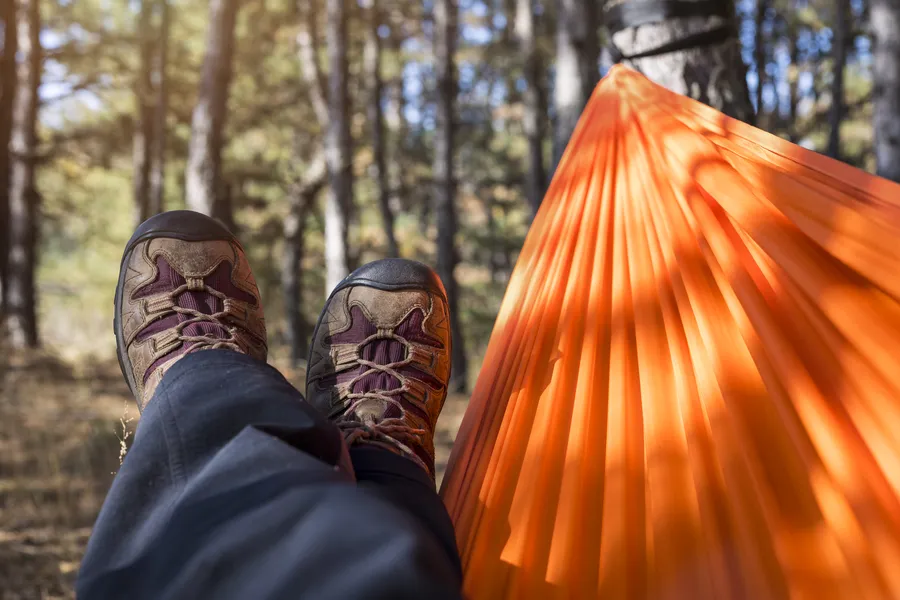 Miehen jalat riippumatossa orange värisessä riippumatossa metsän keskellä, taustalla puita ja luonnon rauhallisuus.