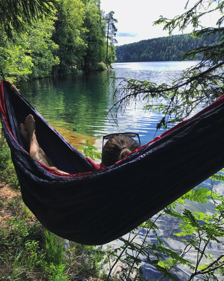 Nuori nainen makaa riippumatossa rauhallisessa maisemassa järven äärellä, nauttien luonnon rauhasta.