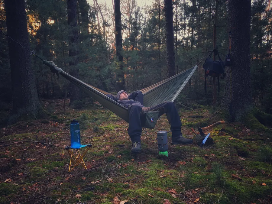 Metsässä oleva mies makaa riippumatossa rentoutuen, vieressä on vesipullo ja keittoastia.
